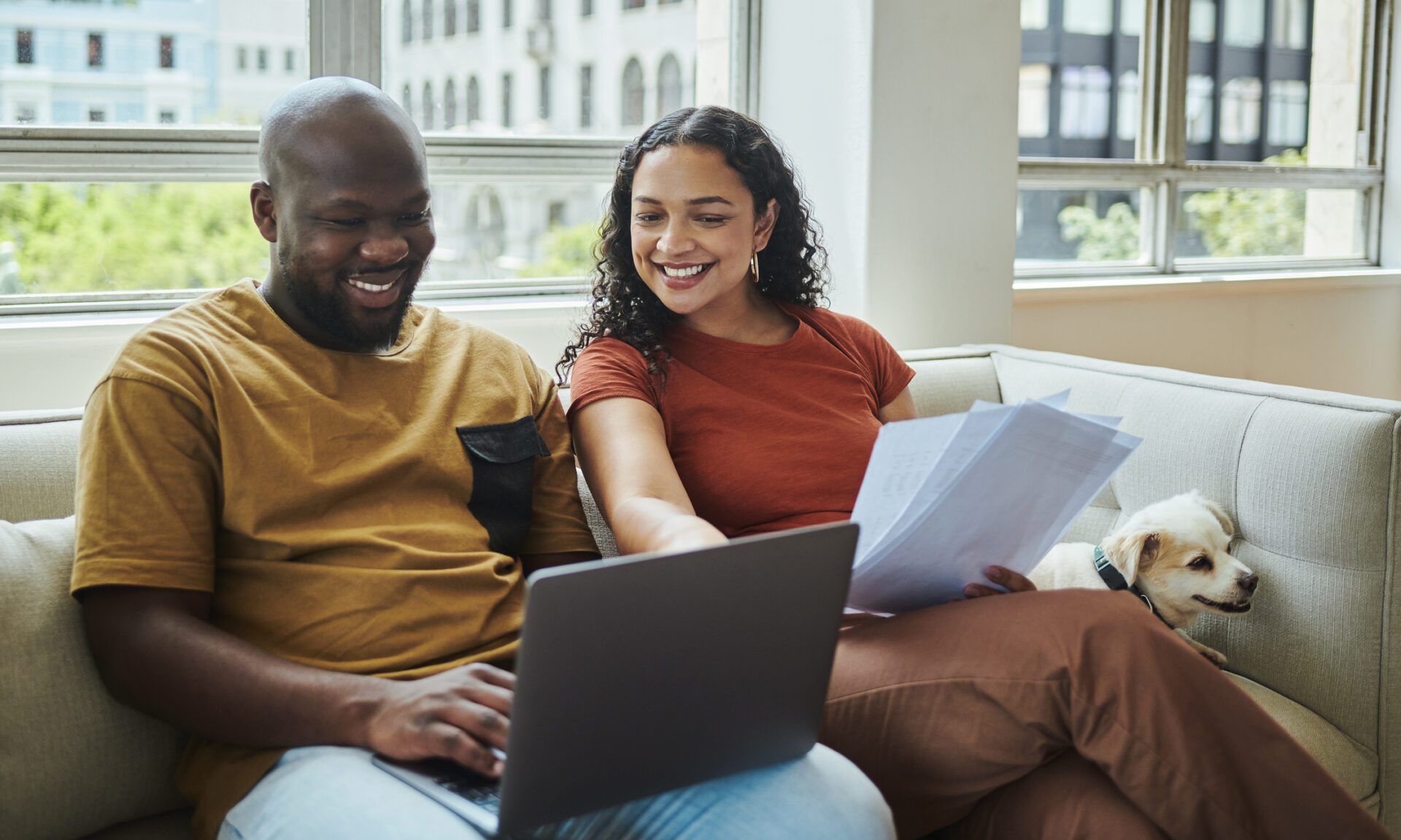A young happy interracial couple working on their budget and finances on a sofa from home with their dog using a laptop computer